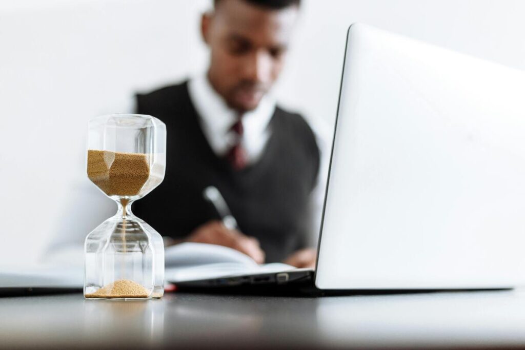 A person is writing at a table with a laptop and an hourglass